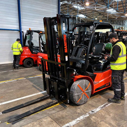 A red and black Manitou forklift is parked inside a warehouse with two workers in high-visibility safety vests standing beside it. The forklift in the foreground has snow socks mounted.