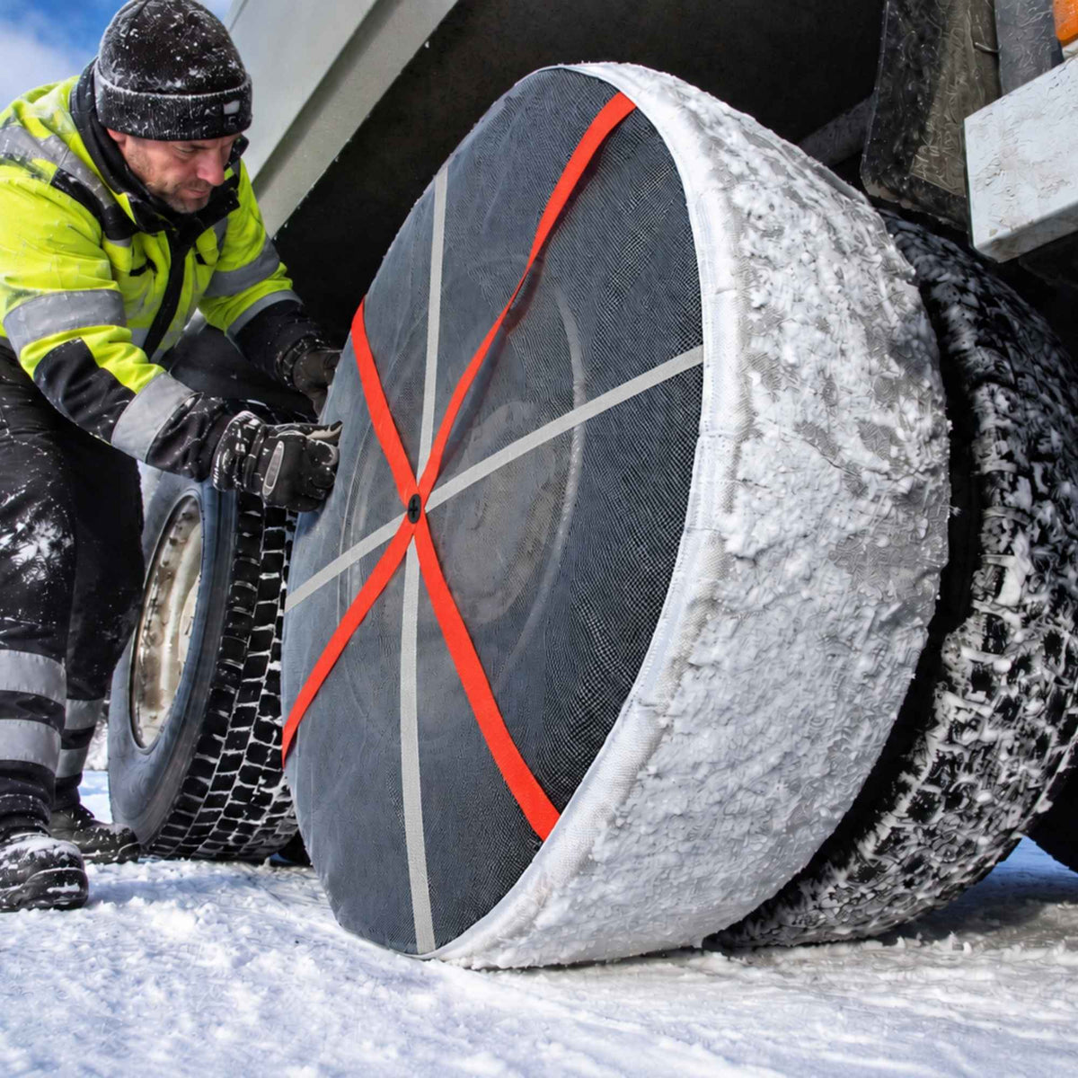 A truck driver in a high-visibility winter jacket is attaching a bright orange strap snow sock to a large winter tire on a truck tire in the snow.