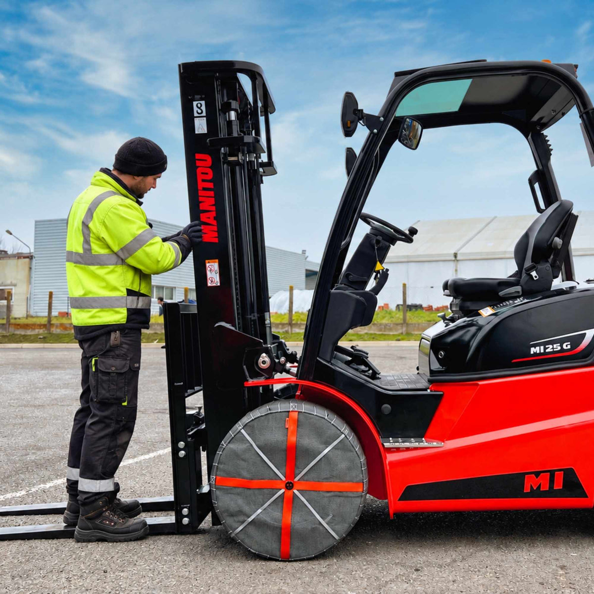 A red and black Manitou forklift parked outdoors on asphalt, with a worker in a high-visibility yellow jacket standing beside the mast and forks under a blue sky. Snow socks are mounted on front wheels.