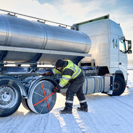 A worker in high-visibility winter clothing stands beside a large silver tanker truck on a snowy road mounting snow sock on rear wheel.