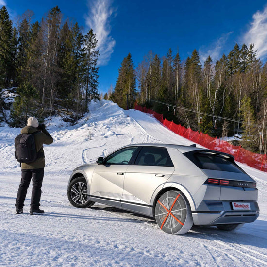 A silver electric crossover SUV is parked on a snowy mountain road and snow chains mounted on rear wheels with a photographer taking a photo beside it. The scene includes evergreen trees, a snow slope, and a red safety fence under a blue sky.